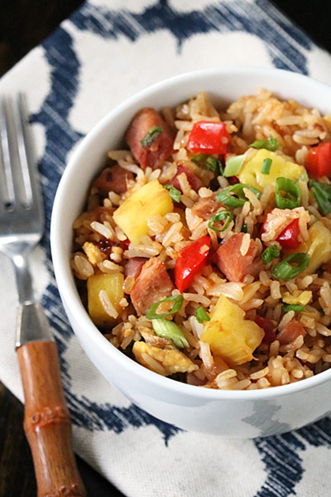 A white bowl filled with fried rice, diced ham, pineapple chunks, red bell peppers, and green onions, placed on a patterned cloth napkin next to a fork with a wooden handle.