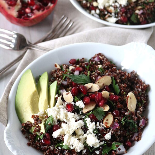 A white bowl filled with quinoa salad topped with feta cheese, pomegranate seeds, sliced almonds, herbs, and avocado slices. Another bowl and a halved pomegranate are in the background on a light table.