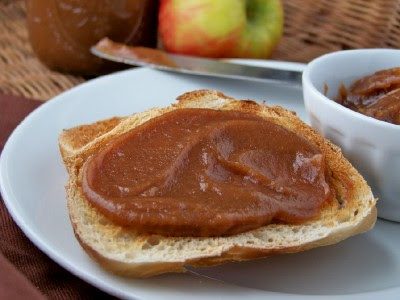 A slice of toasted bread topped with a thick layer of apple butter on a white plate, with a bowl of more apple butter, a knife, and an apple in the background.