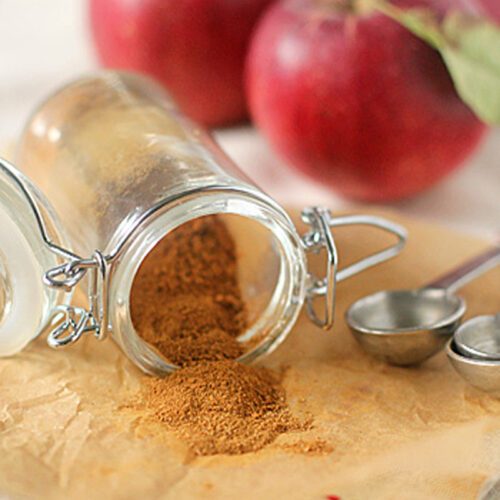 A small glass jar of ground cinnamon is tipped over, spilling onto parchment paper, with metal measuring spoons and three red apples in the background.
