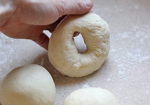 A hand shapes a piece of dough with a hole in the center, forming a bagel. Two more round dough balls rest on a lightly floured surface nearby.