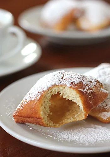A close-up of two powdered sugar-covered beignets on a white plate, one with a large bite taken out, revealing its airy interior. A blurred cup and plate appear in the background.