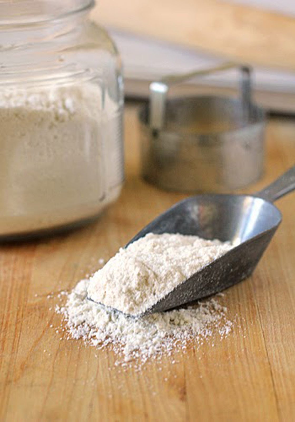 A metal scoop filled with flour rests on a wooden surface, with some flour spilled. A glass jar of flour and a round metal cookie cutter are in the background.