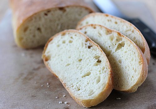 Three slices of freshly baked bread sit on a wooden surface, with the rest of the loaf and a bread knife visible in the background. The bread has a golden crust and a soft, airy crumb.