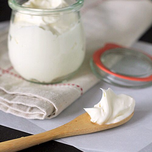 A glass jar filled with creamy white spread sits on a folded cloth with a red stitch, next to a wooden spoon holding a dollop of the spread and an open jar lid on a dark surface.