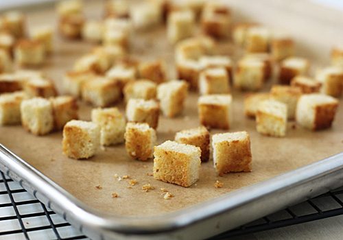 Small, golden-brown bread cubes are spread out on a parchment-lined baking sheet, placed on a wire cooling rack. The bread cubes appear to be homemade croutons.