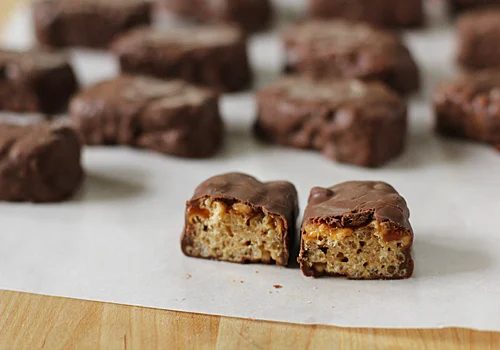 Chocolate-covered cereal bars are arranged on parchment paper, with one bar cut in half at the front, revealing a crispy rice and caramel interior.