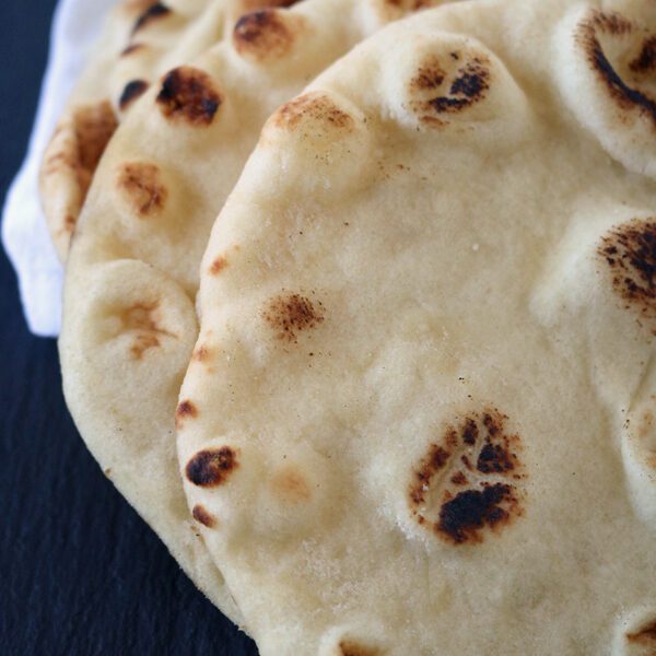 Two pieces of naan bread with golden-brown, slightly charred spots rest partially covered by a white cloth on a dark surface.