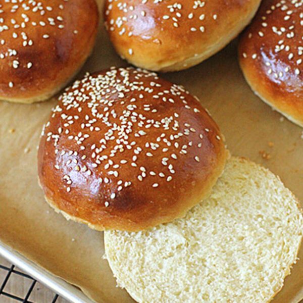 Golden brown sesame seed hamburger buns on a baking tray, with one bun sliced open to show its soft, fluffy interior.