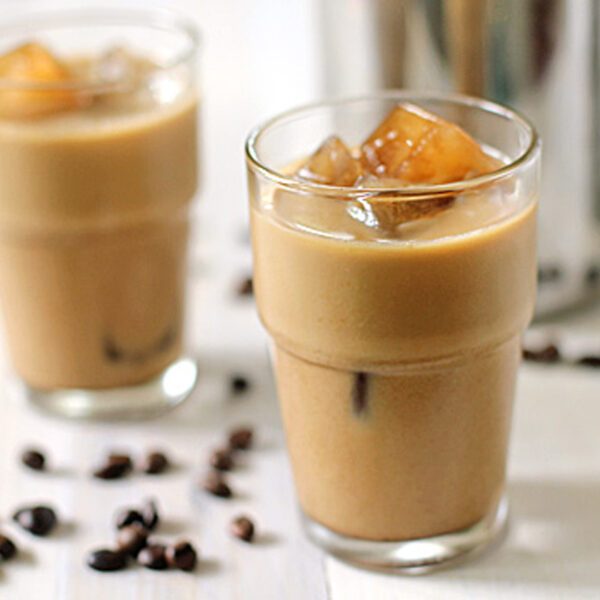 Two glasses of iced coffee with milk, each filled with ice cubes, sit on a white surface. Coffee beans are scattered around the glasses, and a metal pitcher is visible in the background.