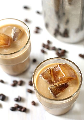 Two glasses of iced coffee with coffee ice cubes, surrounded by scattered coffee beans on a white surface. A metal container is partially visible in the background.