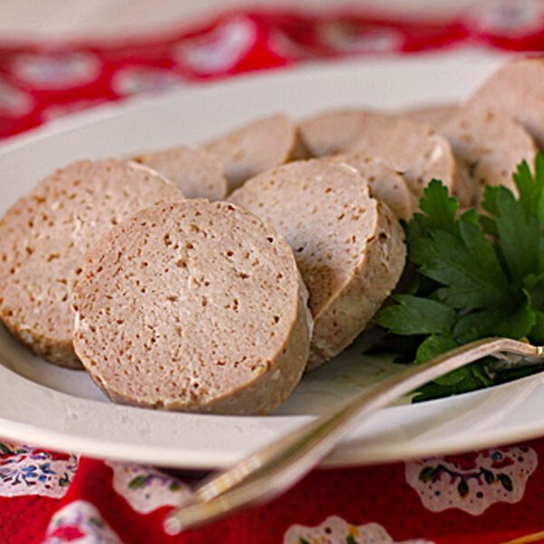 Slices of pale sausage-like meat, possibly a type of luncheon meat or pate, arranged on a white plate with a sprig of parsley and a fork, set on a red patterned cloth.