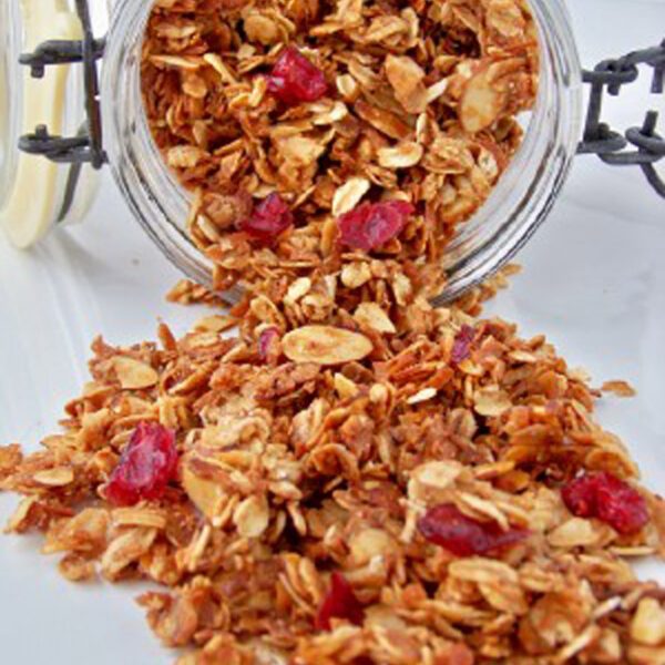 A glass jar tipped over, spilling granola with oats, sliced almonds, and dried cranberries onto a white surface.