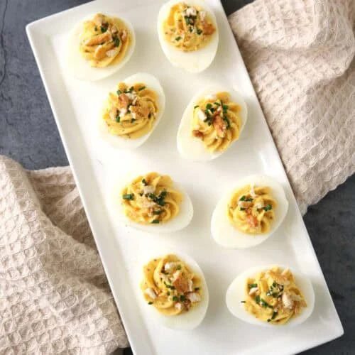 A rectangular white plate with eight Honey-Mustard Deviled Eggs, garnished with herbs and crumbled topping, sits on a gray surface with beige textured towels and a bowl of breadsticks nearby.