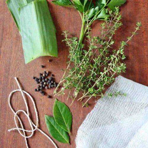 A bunch of herbs, bay leaves, black peppercorns, string, cheesecloth, and leek leaves are arranged on a wooden surface—classic ingredients you’ll need if you’re wondering how to make a bouquet garni and sachet.
