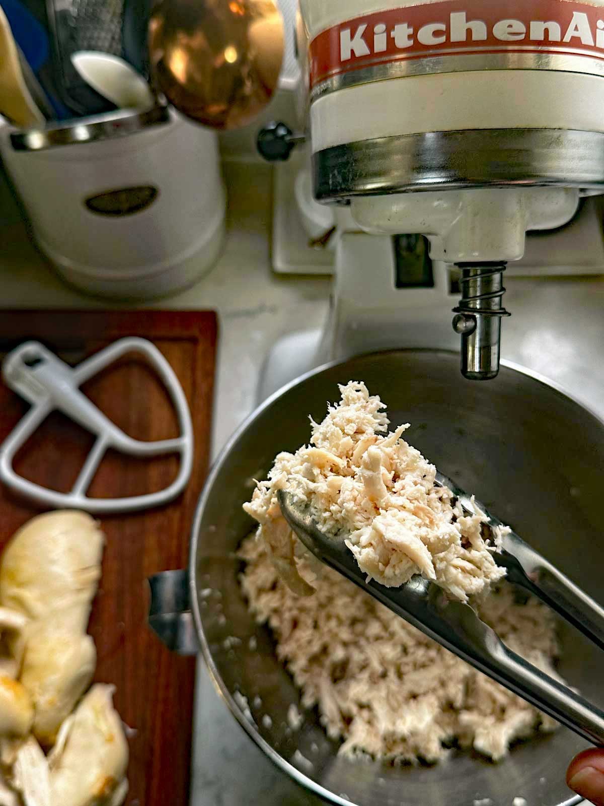 Tongs holding shredded chicken over a mixing bowl attached to a stand mixer, with cooked chicken pieces and a paddle attachment on a cutting board nearby. Kitchen utensils and appliances are in the background.
