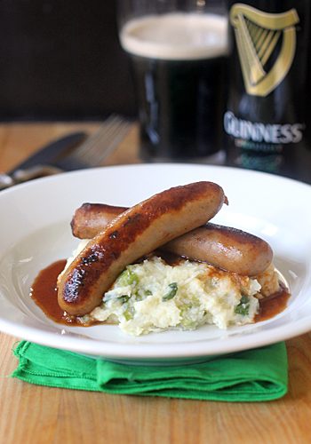 A plate of bangers and mash with gravy, served on a white plate over green napkin. In the background, a glass and bottle of Guinness stout are visible.