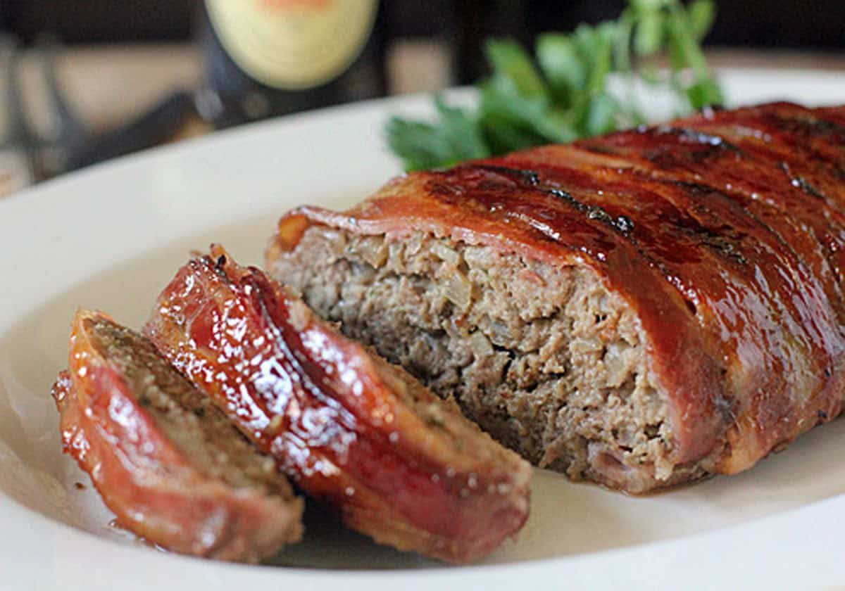 A bacon-wrapped meatloaf is sliced and served on a white plate, with fresh herbs in the background. The meatloaf has a glossy, caramelized glaze on top.