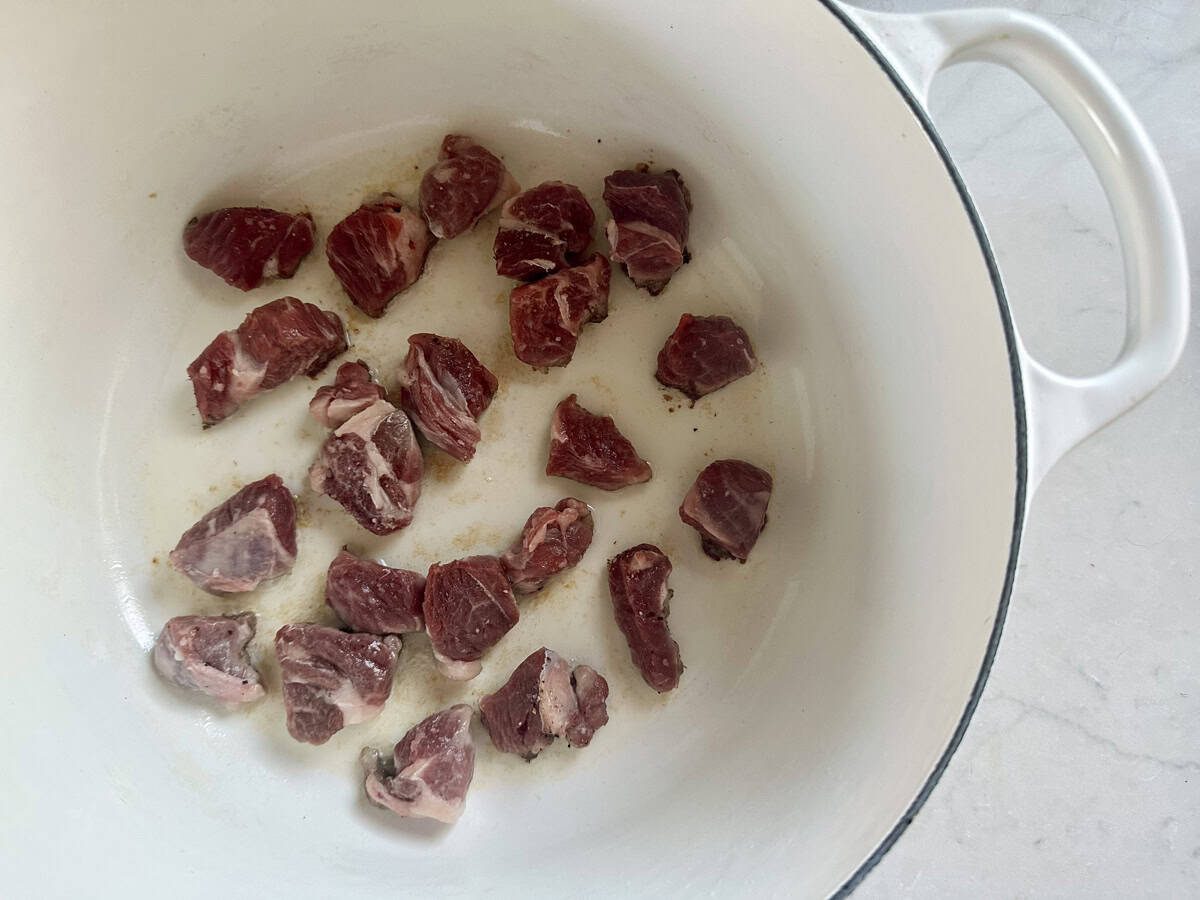 Chunks of raw beef are spread across the bottom of a white enameled pot, ready to be browned, on a light-colored countertop.