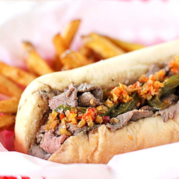 A sandwich filled with sliced beef, peppers, and chopped vegetables, served in a white bun on red-checkered paper, with a side of golden French fries.
