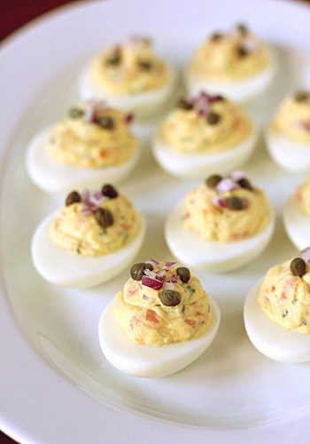 A plate of deviled eggs arranged neatly, each filled with a creamy yellow yolk mixture and topped with capers and finely chopped red onions.