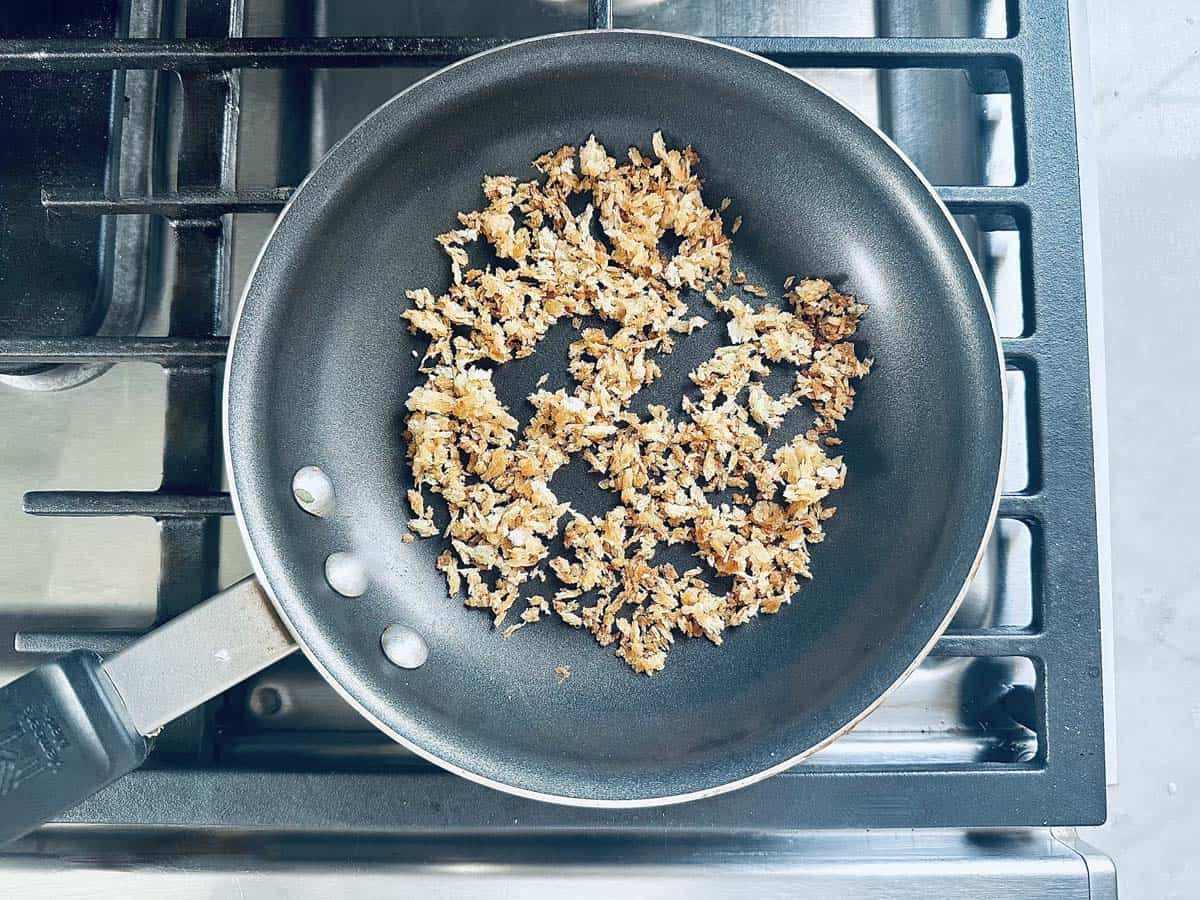 A frying pan on a stove contains browned, crispy shredded coconut, distributed mostly in the center of the pan. The stovetop is metallic with visible burner grates.