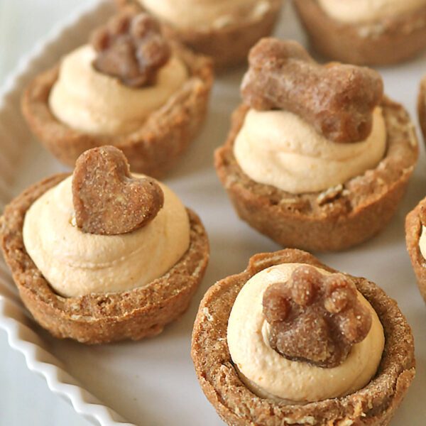 Mini tart-like treats with light tan filling, topped with dog-shaped biscuits, including hearts, bones, and paw prints, arranged on a white plate.