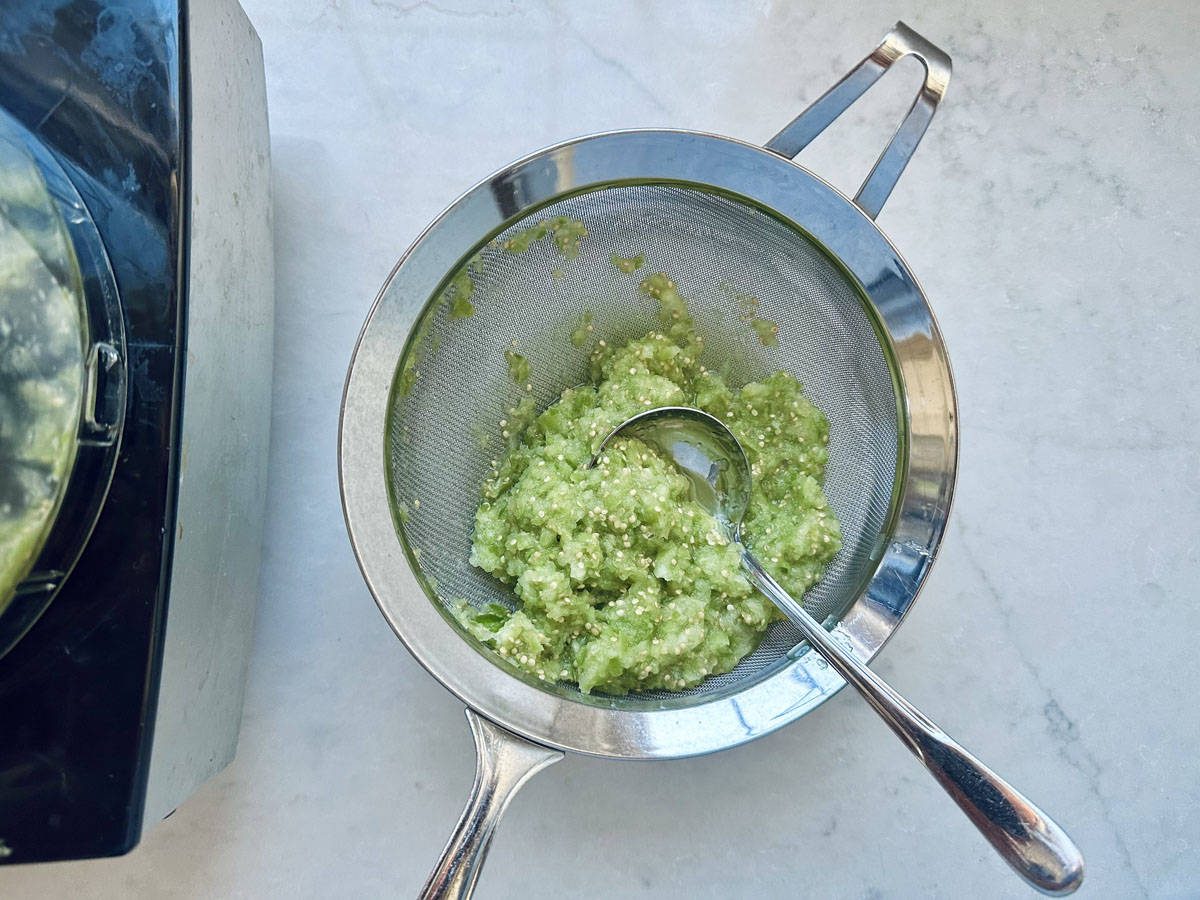A metal strainer containing blended green mixture sits over a bowl on a white countertop, with a spoon resting inside. A food processor is partially visible on the left.