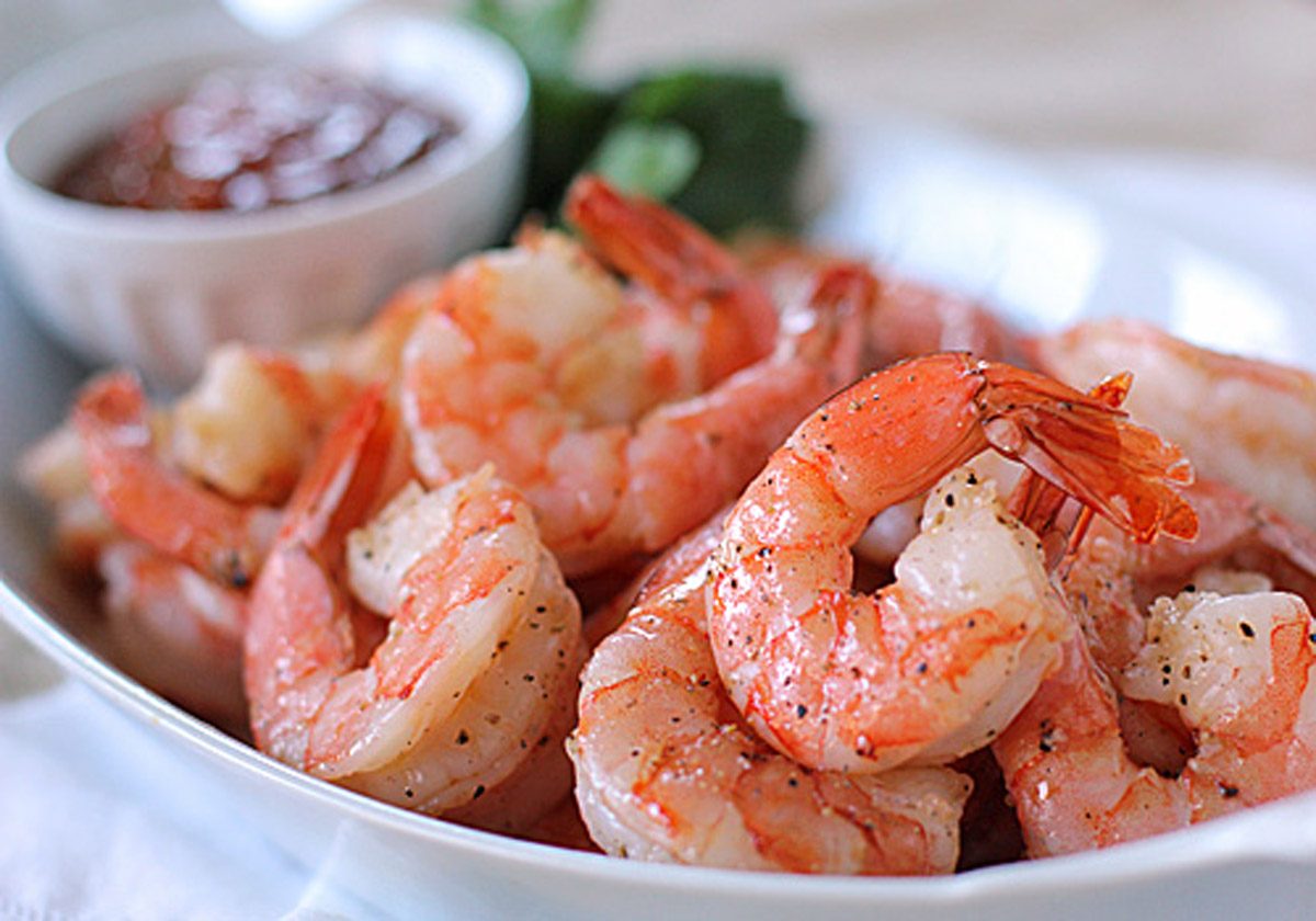 A plate of cooked, seasoned shrimp is served with a small bowl of cocktail sauce in the background. The shrimp are pink, plump, and garnished with ground pepper.