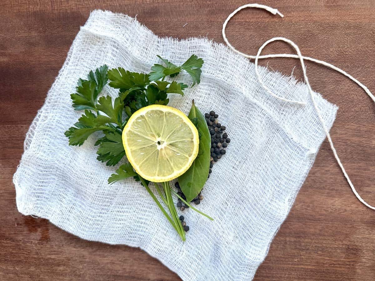 A slice of lemon, parsley, bay leaf, green herbs, and whole black peppercorns arranged on a piece of white cheesecloth, with a string beside them, all on a wooden surface.