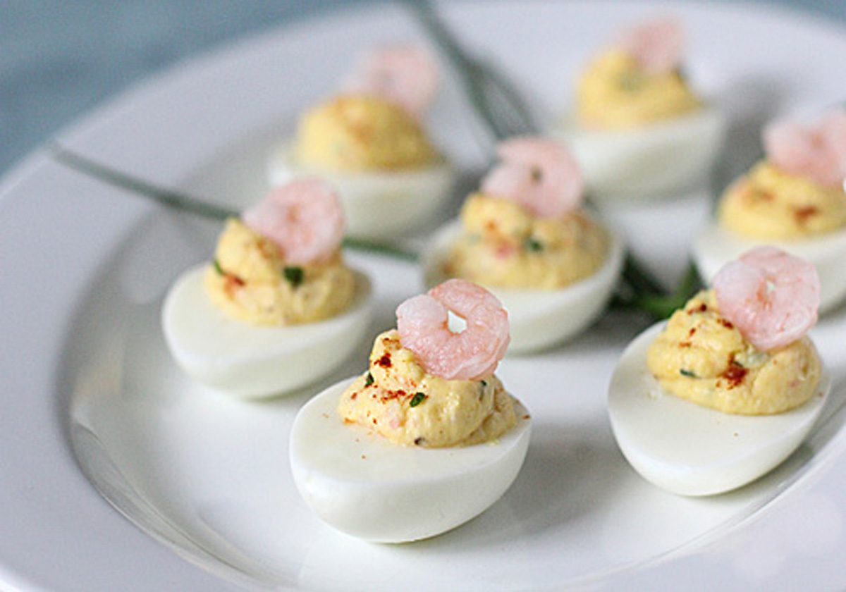 A white plate with several deviled eggs, each topped with a small shrimp and garnished with herbs. The eggs are neatly arranged, and there are some green chives in the background.
