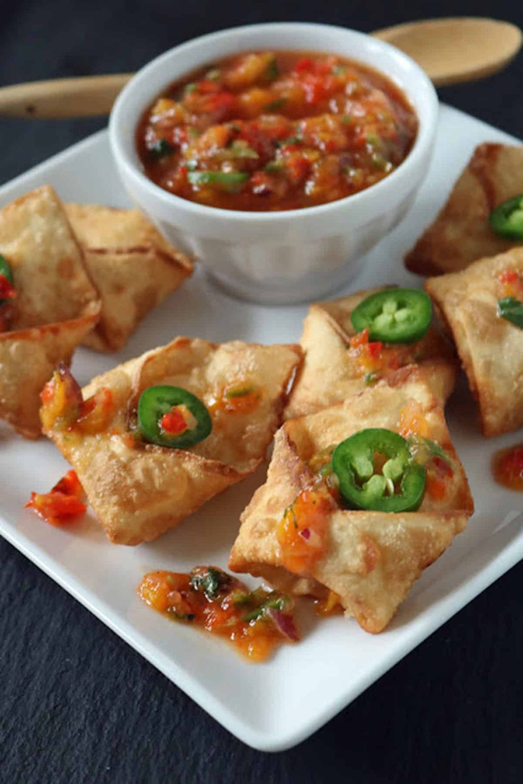 A white plate with crispy wontons topped with sliced jalapeños and sweet chili sauce, served with a bowl of chunky salsa or dip, on a dark surface.