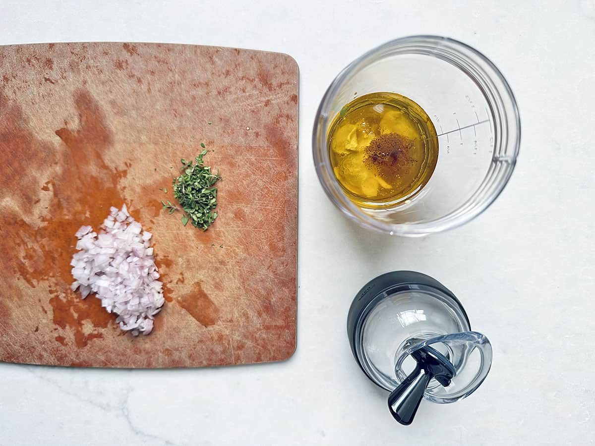 Chopped shallots and herbs on a wooden cutting board next to a blender cup filled with oil, vinegar, and seasoning—perfect for preparing the dressing that will elevate your Kale Apple Harvest Salad—sit alongside a blender lid on a white surface.