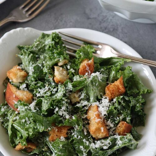 A white bowl filled with kale Caesar salad topped with croutons and grated cheese, with a fork resting inside. Another bowl and a fork are visible in the background on a gray surface.