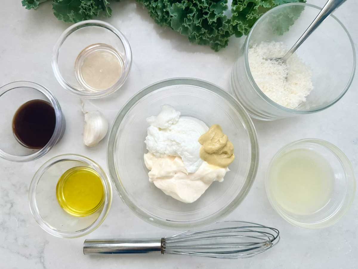 Various ingredients in glass bowls are arranged on a white surface, including mayonnaise, mustard, olive oil, lemon juice, grated cheese, a garlic clove, and leafy greens. A whisk lies nearby.