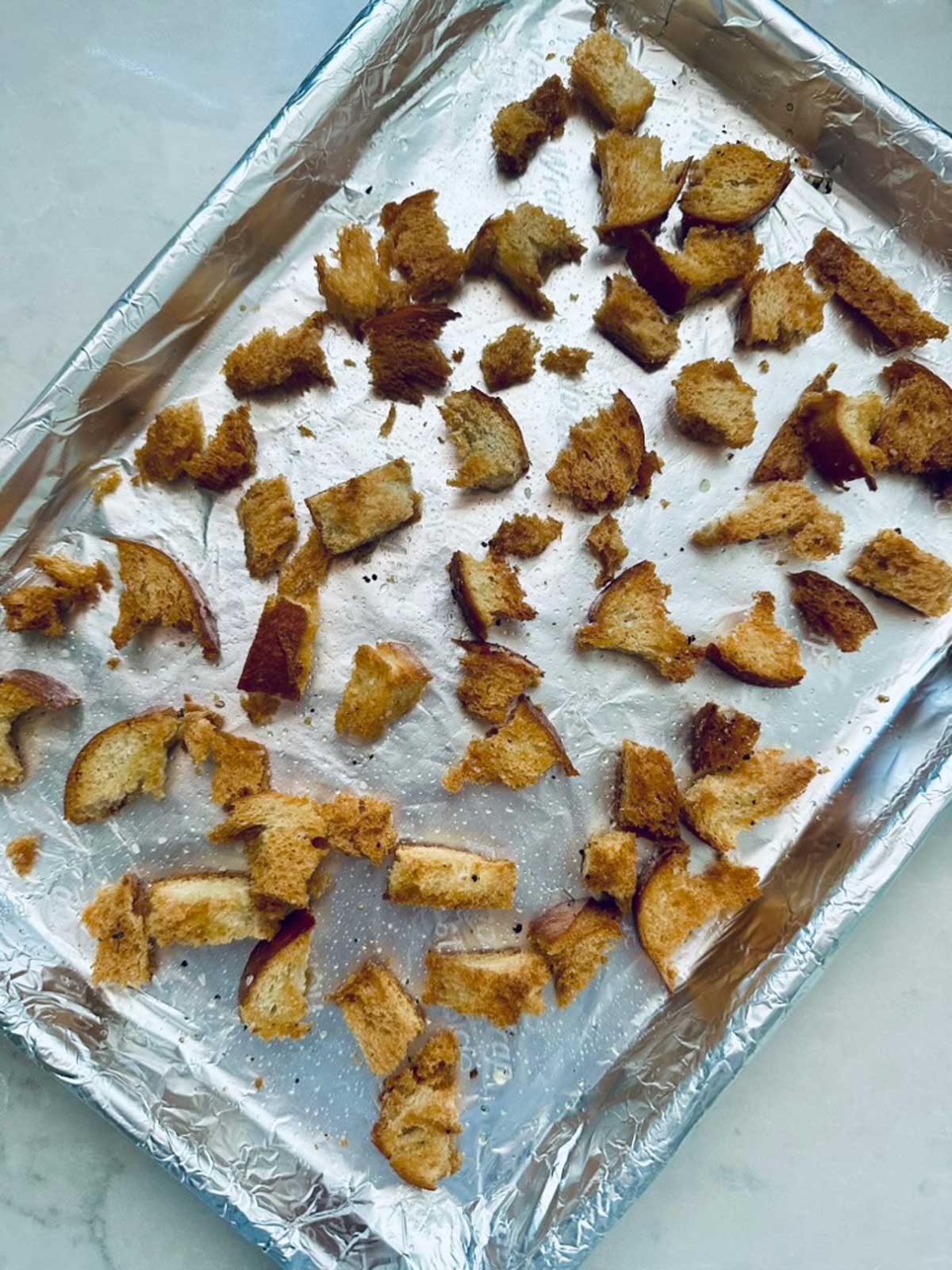 Cubed pieces of toasted bread are scattered on a foil-lined baking sheet, likely prepared as homemade croutons. The bread pieces are golden brown and uneven in size.