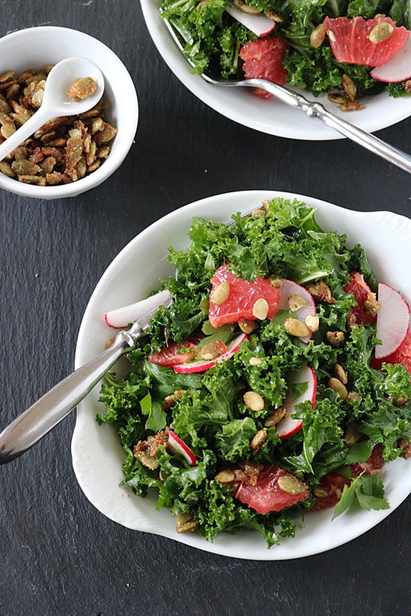 A bowl of kale salad with sliced radishes, grapefruit segments, and crunchy seeds. A spoon rests in the bowl. Another smaller white dish with seeds and a spoon sits nearby on a dark surface.