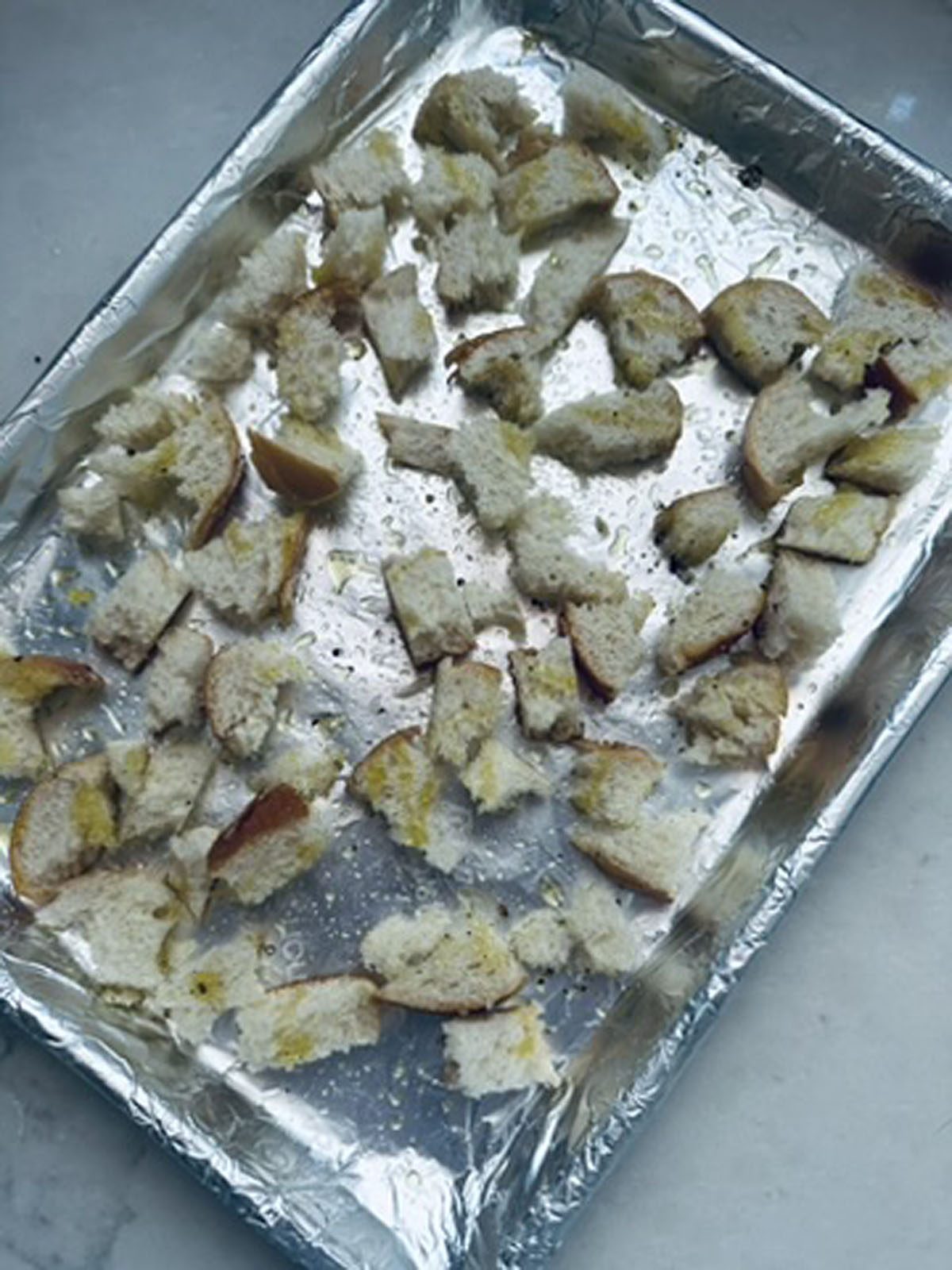 A foil-lined baking sheet with scattered pieces of torn bread drizzled with oil and sprinkled with seasoning, ready to be toasted into croutons.