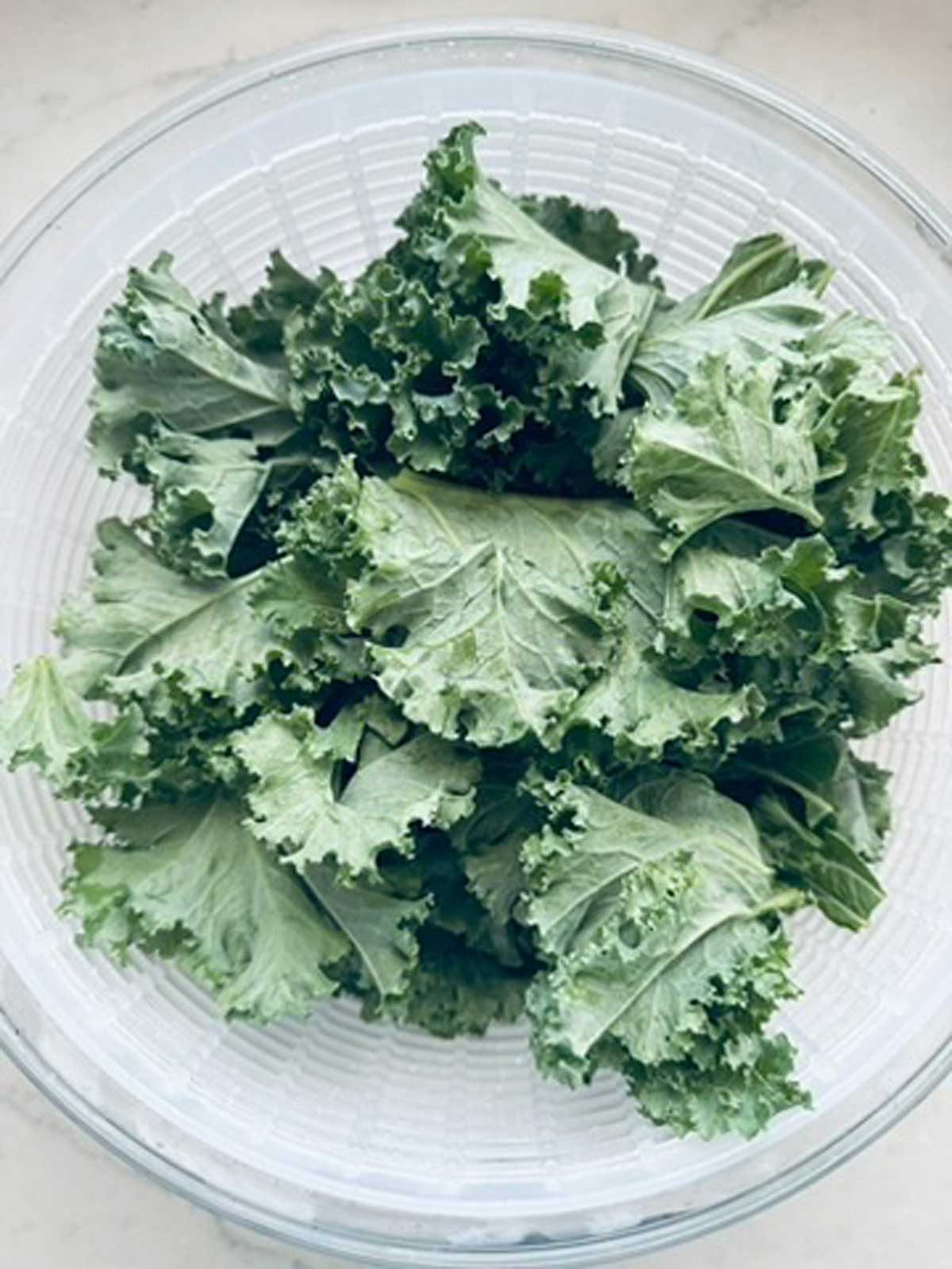 A clear bowl filled with fresh, curly green kale leaves sits on a light-colored surface.