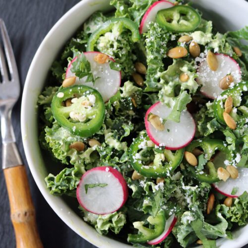 A bowl of kale salad topped with sliced radishes, jalapeños, pepitas, crumbled cheese, and fresh herbs is next to a fork with a wooden handle on a dark surface.