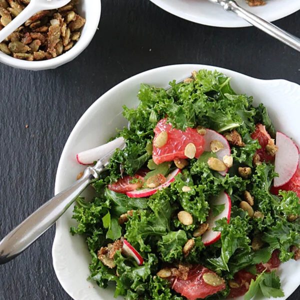 A bowl of kale salad with sliced radishes, grapefruit segments, and crunchy seeds. A spoon rests in the bowl. Another smaller white dish with seeds and a spoon sits nearby on a dark surface.