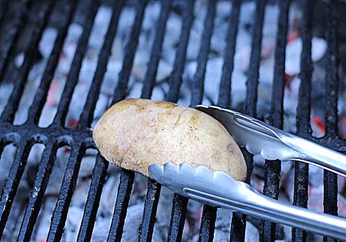 A metal tong is holding a whole potato on a grill over hot coals, preparing it for cooking.