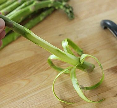 A hand holds a stalk of asparagus with its lower portion peeled. Thin strips of asparagus skin curl around the stalk. A vegetable peeler and more asparagus stalks are on a wooden surface in the background.