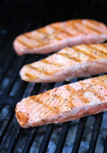 Three seasoned salmon fillets are cooking on a grill, showing visible grill marks. The fillets are arranged in a row, and the grill grates are blackened from use.