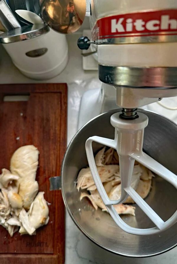 A stand mixer with shredded chicken and a paddle attachment sits on a counter beside a cutting board with more cooked chicken being torn into pieces. Kitchen utensils are in the background.