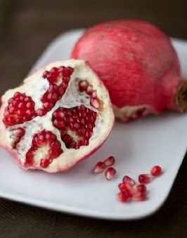 A whole pomegranate and a half-open pomegranate with bright red seeds are displayed on a white plate. Several loose seeds are scattered on the plate near the fruit.