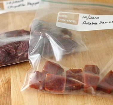Two labeled resealable plastic bags on a wooden surface; one contains dried chili peppers, the other contains small frozen cubes of adobo sauce. Handwritten labels indicate the contents and date.