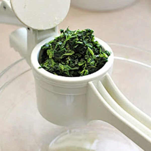 A white handheld potato ricer filled with chopped spinach is positioned over a clear glass bowl, ready to press the spinach through.