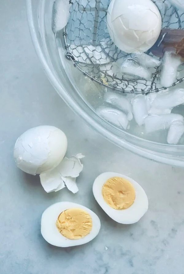Peeled hard-boiled eggs on a marble surface, one sliced in half to show the yolk, next to a bowl of ice water with eggs and a slotted spoon inside.