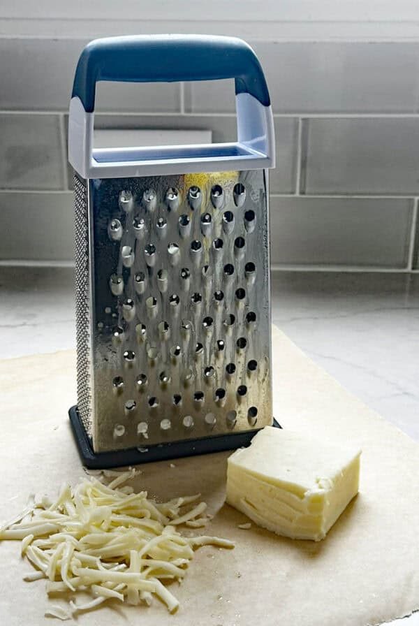 A box cheese grater stands on parchment paper next to a block of cheese and a small pile of grated cheese, with a tiled backsplash in the background.